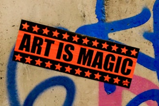 A vibrant sticker with the text 'Art is Magic' is plastered on a rough concrete wall. The sticker is orange with black stars framing bold black letters. The wall features graffiti in blue and pink spray paint, creating an urban, artistic backdrop.