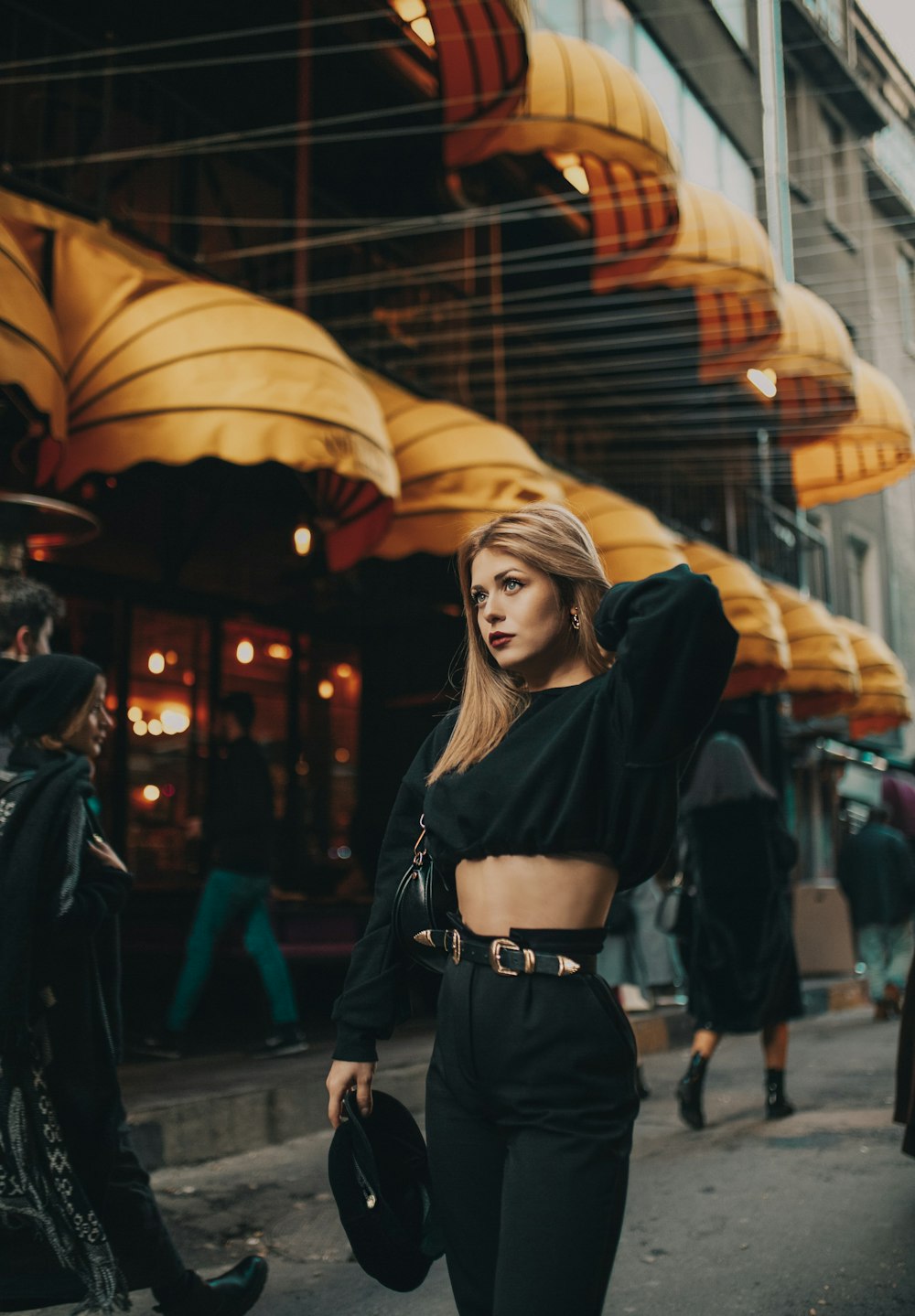 woman wearing black crew-neck sweatshirt holding black hat while standing on pathway near people walking beside building during daytime