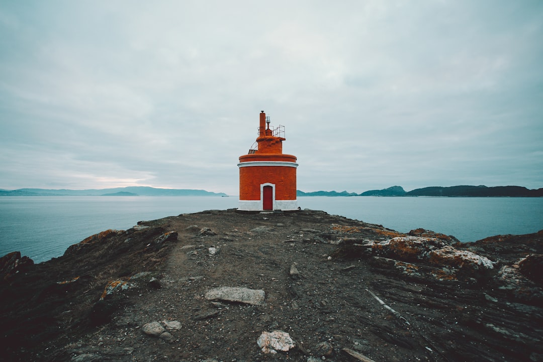 orange and white lighthouse o cliff viewing blue body of water under white and blue sky