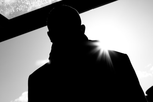 A silhouette of a person is seen against a bright light source, creating a dramatic contrast. The light produces a lens flare effect, and the scene has a high contrast with deep shadows.