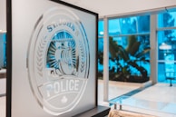An indoor scene showing a frosted glass panel with an etched design of a seal featuring a large ship and the words 'ST. JOHN'S POLICE'. The background is a modern interior with large windows and tropical plants, illuminated with a cool blue light.
