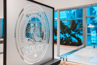 An indoor scene showing a frosted glass panel with an etched design of a seal featuring a large ship and the words 'ST. JOHN'S POLICE'. The background is a modern interior with large windows and tropical plants, illuminated with a cool blue light.