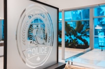 An indoor scene showing a frosted glass panel with an etched design of a seal featuring a large ship and the words 'ST. JOHN'S POLICE'. The background is a modern interior with large windows and tropical plants, illuminated with a cool blue light.