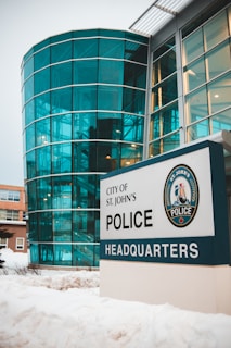 A modern building with large glass windows and a sign in front that reads 'City of St. John's Police Headquarters'. The building features a teal-tinted, semi-circular glass facade and is surrounded by snow.
