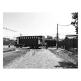 A large truck is parked on a dirt path surrounded by fencing and utility poles. There is a person guiding a horse towards the truck and another person walking nearby. The scene appears to be outdoors in a semi-industrial area with visible power lines and some vegetation in the background.