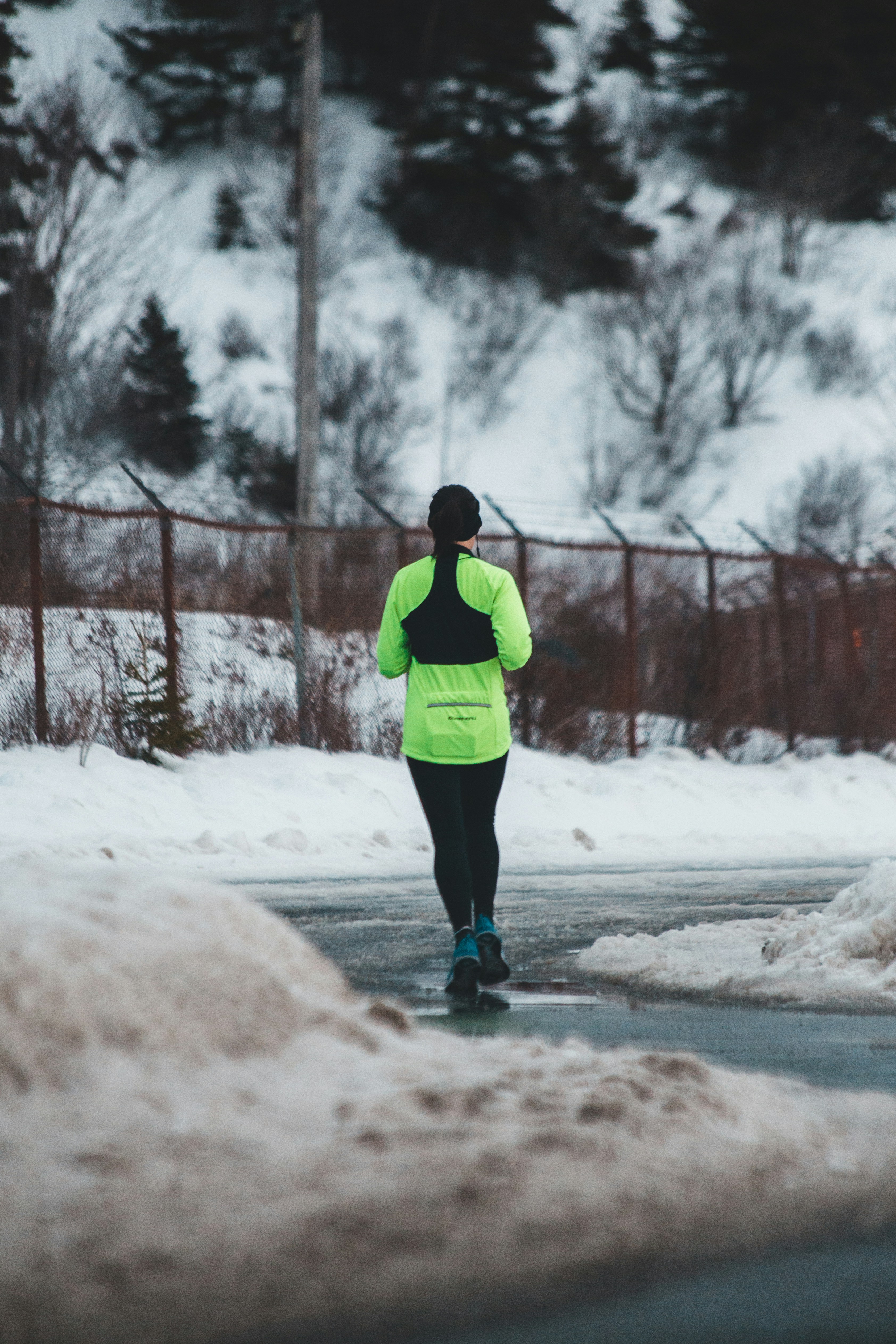 Runner seen from behind in a bright green jacket and black leggings on a snowy road beside a fence