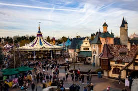 A bustling amusement park setting features a colorful carousel with a pointed roof adorned with flags. Surrounding the carousel, there are cobblestone pathways lined with quaint, fantasy-style buildings with ornamental facades and varied roof designs. Numerous visitors wander the area, some pushing strollers, and others standing in groups or walking leisurely. Trees and manicured bushes add to the whimsical landscape.