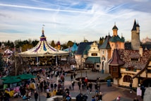A bustling amusement park setting features a colorful carousel with a pointed roof adorned with flags. Surrounding the carousel, there are cobblestone pathways lined with quaint, fantasy-style buildings with ornamental facades and varied roof designs. Numerous visitors wander the area, some pushing strollers, and others standing in groups or walking leisurely. Trees and manicured bushes add to the whimsical landscape.