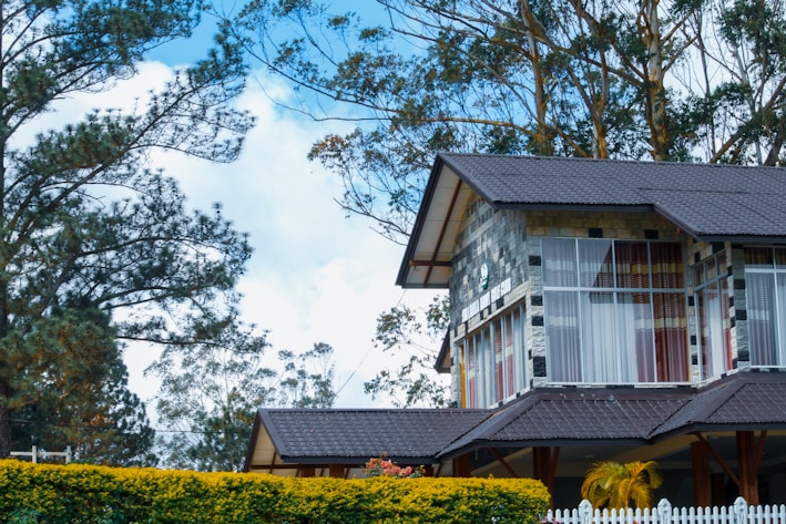A house with a modern design features large glass windows and a sloping roof made of dark shingles. The exterior is composed of a combination of stone and wood elements. Surrounding the house are lush trees and shrubs, adding a natural backdrop. A white picket fence is visible in the foreground.
