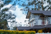 A house with a modern design features large glass windows and a sloping roof made of dark shingles. The exterior is composed of a combination of stone and wood elements. Surrounding the house are lush trees and shrubs, adding a natural backdrop. A white picket fence is visible in the foreground.