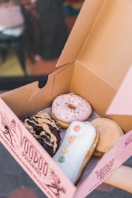 Inside a pink box from Voodoo Doughnut, four doughnuts are displayed. One doughnut has pink icing with colorful sprinkles, another is chocolate with peanut butter drizzle, a third doughnut has white icing with colorful loops resembling a caterpillar, and the last one is sugar-coated.