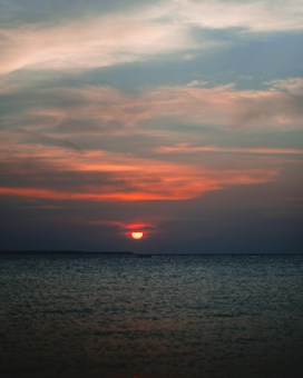 The image captures a serene sunset over a calm sea, with a vivid orange sun descending towards the horizon. The sky is painted with soft hues of pink, orange, and blue, reflecting beautifully on the water's surface.