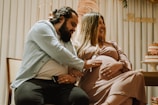 A couple is sitting together in a warmly decorated room, with the man gently placing his hand on the woman's pregnant belly. Both are smiling affectionately, creating a heartwarming and intimate atmosphere.