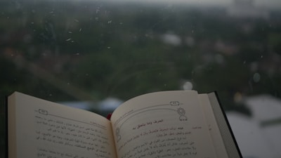 A book open to a page with Arabic text, positioned near a window with an out-of-focus view of green foliage and overcast sky in the background.