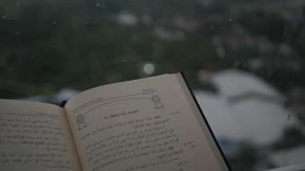 An open book is held up with text written in Arabic. The page is adorned with decorative elements. The background through a window is blurred, showing a green landscape with some structures.
