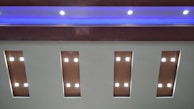 A ceiling with a modern design features recessed wooden panels, each containing an arrangement of bright, white square lights. A blue ambient light runs along the upper section, adding a sleek, contemporary feel.
