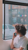 A person with curly hair is sitting near a window, wearing a white shirt and holding a phone to their ear. Outside the window, there is a view of a street with a parked white car and a storefront with a colorful pizza sign.