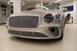 A luxury car with a sleek, metallic exterior and distinctive grille is displayed inside a modern showroom. The lighting highlights the vehicle's glossy finish and elegant design features.