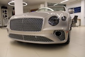 A luxury car with a sleek, metallic exterior and distinctive grille is displayed inside a modern showroom. The lighting highlights the vehicle's glossy finish and elegant design features.