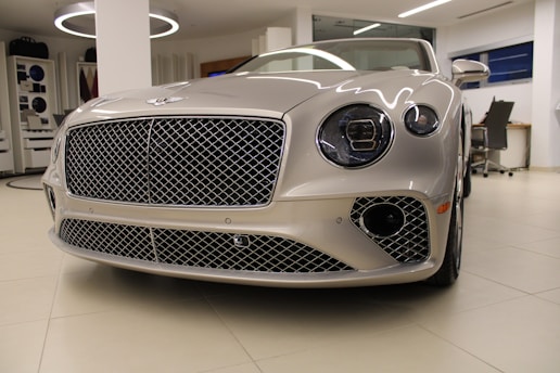 A luxury car with a sleek, metallic exterior and distinctive grille is displayed inside a modern showroom. The lighting highlights the vehicle's glossy finish and elegant design features.