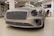 A luxury car with a sleek, metallic exterior and distinctive grille is displayed inside a modern showroom. The lighting highlights the vehicle's glossy finish and elegant design features.