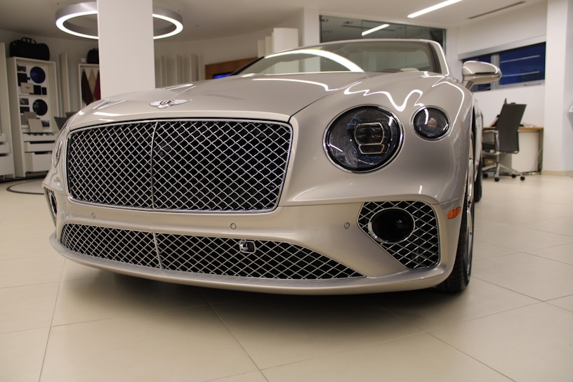 A luxury car with a sleek, metallic exterior and distinctive grille is displayed inside a modern showroom. The lighting highlights the vehicle's glossy finish and elegant design features.