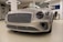 A luxury car with a sleek, metallic exterior and distinctive grille is displayed inside a modern showroom. The lighting highlights the vehicle's glossy finish and elegant design features.