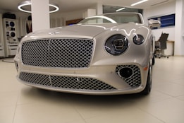 A luxury car with a sleek, metallic exterior and distinctive grille is displayed inside a modern showroom. The lighting highlights the vehicle's glossy finish and elegant design features.