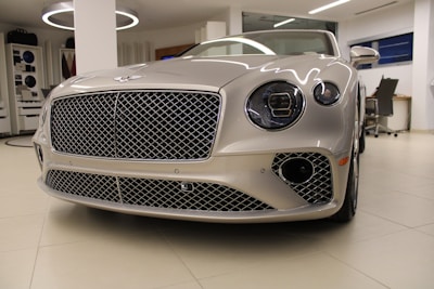 A luxury car with a sleek, metallic exterior and distinctive grille is displayed inside a modern showroom. The lighting highlights the vehicle's glossy finish and elegant design features.