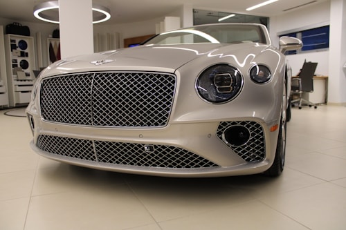 A luxury car with a sleek, metallic exterior and distinctive grille is displayed inside a modern showroom. The lighting highlights the vehicle's glossy finish and elegant design features.