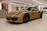A sleek, luxurious sports car with a golden-yellow finish is parked indoors on a polished tile floor. The car features distinctive black rims and aerodynamic curves. The setting is modern and well-lit, with wooden panels and white walls in the background.