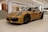 A sleek, luxurious sports car with a golden-yellow finish is parked indoors on a polished tile floor. The car features distinctive black rims and aerodynamic curves. The setting is modern and well-lit, with wooden panels and white walls in the background.