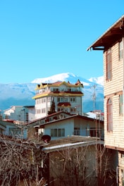 A vibrant scene featuring a unique multi-story building with yellow accents on the roof, surrounded by residential houses. Behind the structures, snow-capped mountains rise majestically against a bright blue sky. The image suggests a mix of urban and natural elements, with leafless tree branches in the foreground and satellite dishes visible on the rooftops.