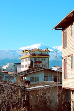 A vibrant scene featuring a unique multi-story building with yellow accents on the roof, surrounded by residential houses. Behind the structures, snow-capped mountains rise majestically against a bright blue sky. The image suggests a mix of urban and natural elements, with leafless tree branches in the foreground and satellite dishes visible on the rooftops.