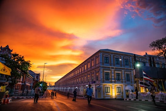 The image captures a vibrant sunset with intense orange and yellow hues illuminating the sky. A grand building with classic architecture stretches along the street as people walk leisurely. Flags adorn the area, while streetlights add a modern touch to the historical setting.