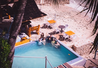 A group of people enjoy a swim in a small pool located by a rocky beach. The setting includes wooden seating and tables with orange umbrellas. The surrounding area is a mix of stone paths and sandy shore, framed by palm leaves.