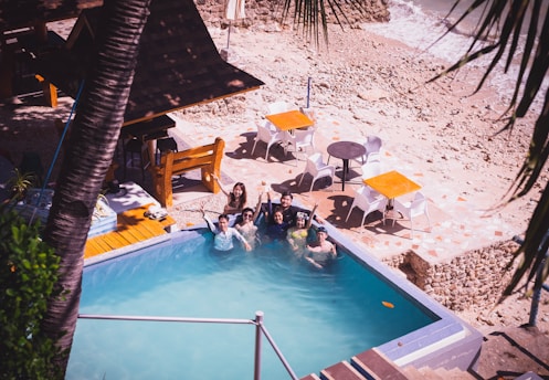 A group of people enjoy a swim in a small pool located by a rocky beach. The setting includes wooden seating and tables with orange umbrellas. The surrounding area is a mix of stone paths and sandy shore, framed by palm leaves.