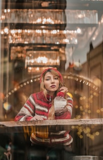 A person wearing a cozy, striped red and beige sweater is sitting inside, with soft lighting and decorative light fixtures creating a warm, inviting atmosphere. They appear thoughtful, resting their chin on one hand while looking outside.