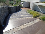 A stone pathway integrates seamlessly into a landscaped garden, bordered by robust stone walls. Nearby greenery and small trees complement the natural materials used in the modern architectural design of the nearby building with its light stone facade.