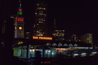 Nighttime cityscape featuring a brightly lit clock tower with a colorful facade and an illuminated 'San Francisco' sign. The skyline in the background is composed of tall buildings with lights scattered across windows.