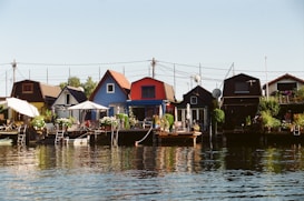 A row of colorful waterfront houses with small porches and lush greenery. The buildings are painted in various tones, including blue, red, and brown. Several umbrellas offer shade, and there are potted plants placed around the area. The calm water reflects the vibrant colors of the structures and the sky is clear.