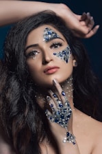 A person with long dark hair features intricate jewelry designs on their face and hand. The decorations include blue and silver stones, adding an elaborate, artistic element. The hand is positioned delicately under the chin, and the gaze is directed upward, creating a dynamic composition.