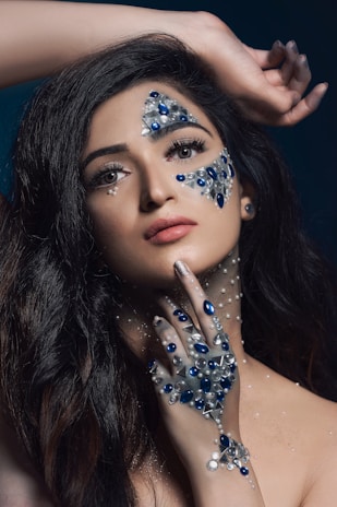 A person with long dark hair features intricate jewelry designs on their face and hand. The decorations include blue and silver stones, adding an elaborate, artistic element. The hand is positioned delicately under the chin, and the gaze is directed upward, creating a dynamic composition.