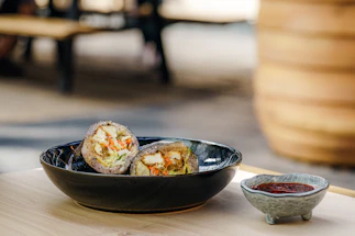 A black bowl contains a sliced food item filled with vegetables and possibly tofu. Beside the bowl, a small gray ceramic dish holds a red sauce. Both items are placed on a light wooden surface with an out-of-focus outdoor setting in the background.