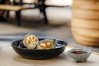 A black bowl contains a sliced food item filled with vegetables and possibly tofu. Beside the bowl, a small gray ceramic dish holds a red sauce. Both items are placed on a light wooden surface with an out-of-focus outdoor setting in the background.