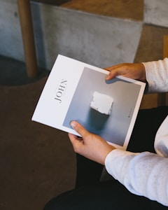 A person is holding a booklet with the title 'JOHN' visible on the cover. The cover features an image of a white object on a light gray background. The person is wearing a long-sleeve, light-colored shirt and is seated indoors on a dark surface.