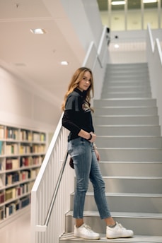 A young woman stands confidently on a modern, minimalistic staircase. She is wearing a black long-sleeve top, blue jeans, and white sneakers. The setting appears to be a library, with shelves filled with colorful books lined along the wall behind her.