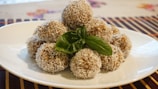 A neatly arranged pile of coconut-covered dessert balls is presented on a white plate. A fresh green basil leaf is placed on top, adding a touch of color. The background features a striped table mat.