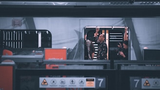 A train station scene with a train stopped on the tracks. Several passengers are visible standing inside the train, holding onto overhead handles. The exterior of the train displays advertisements, and the platform is marked with safety signs and notices.