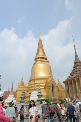 A large golden stupa surrounded by ornate structures with intricate details stands prominently against a partly cloudy sky. The architecture features spires and decorative elements that are characteristic of traditional Southeast Asian temple design. Numerous people are gathered around, some taking photos, indicative of a popular tourist destination.
